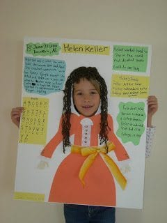 Young student peeking her face through a cut out in a poster sized report on Helen Keller 