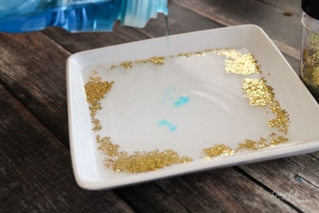 Square dish filled with water and glitter, showing how a drop of dish soap repels the glitter