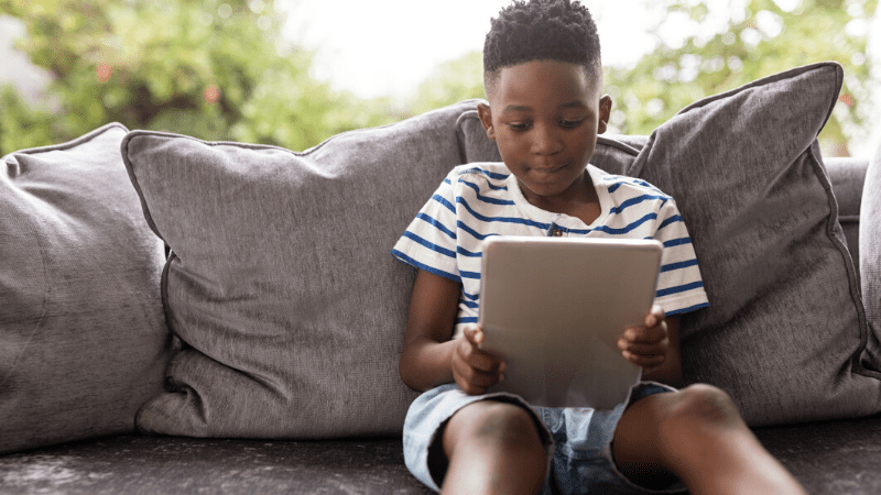 young boy sitting on sofa reading on tablet - teaching reading remotely