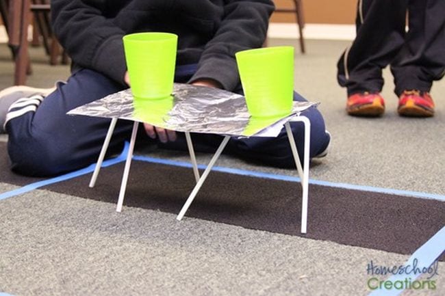 Student showing a foil platform balanced on drinking straws, holding two yellow plastic cups of water