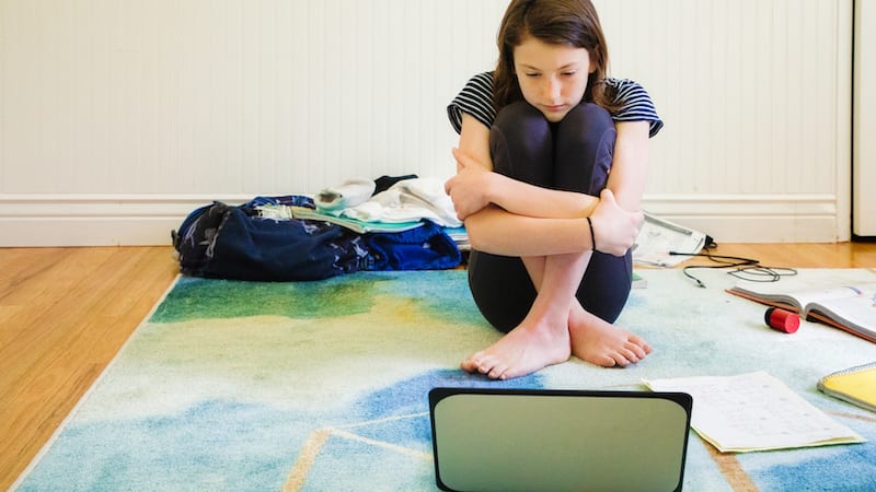 Sad female teenager watching media on laptop hugging herself sitting on the carpet with homework material and clothing around her.