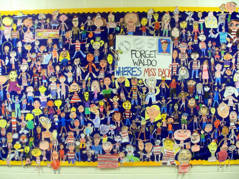 Bulletin board covered in cut-out illustrations of students, with a sign saying "Forget Waldo: Where's Miss Back?"