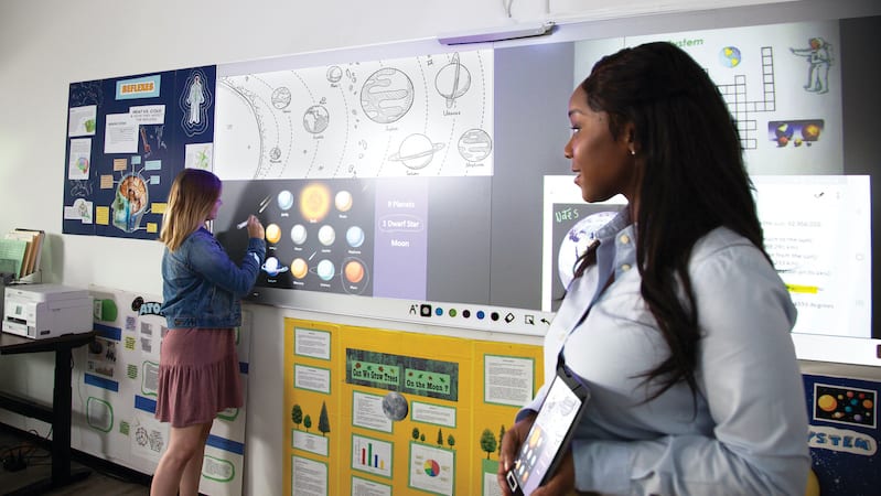 A whiteboard filled of posters of planets and the teacher holding up a tablet with planets.