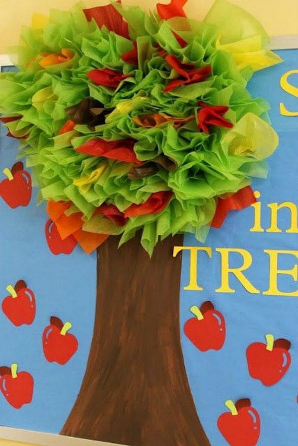 A fall tree is surrounded by apples. The leaves are created from bunched up tissue paper in red, orange, yellow, and green.