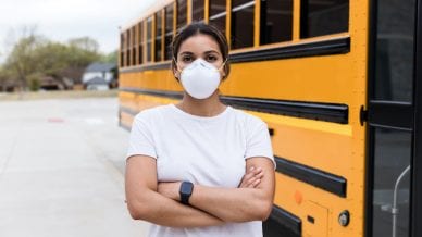 A female school bus driver, wearing an N95 mask, stands next to a school bus. She is standing with her arms crosses and is looking at the camera.