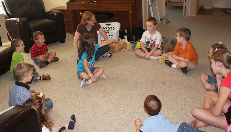 Children sit in a circle on a rug with another child inside the circle. The girl in the circle is pointing toward one of the children on the outside.