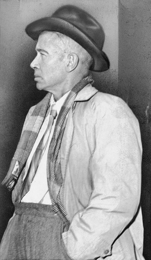 Black and white side view photograph of EE Cummings wearing jacket and hat with hands in pockets.