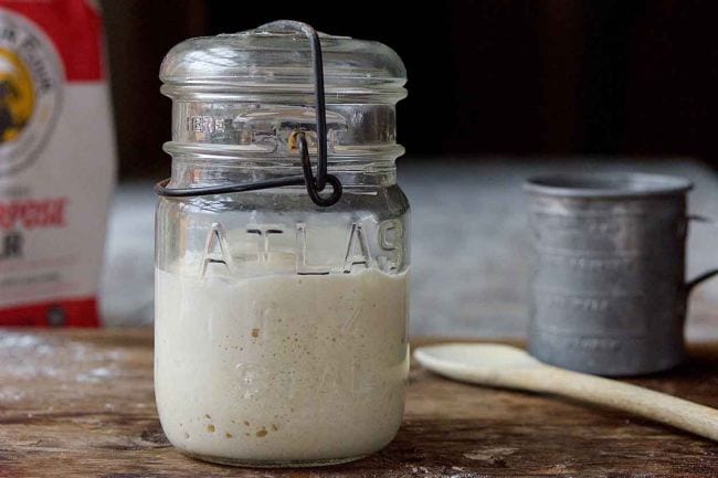 Jar containing sourdough starter, with flour bag, tin cup, and wooden spoon