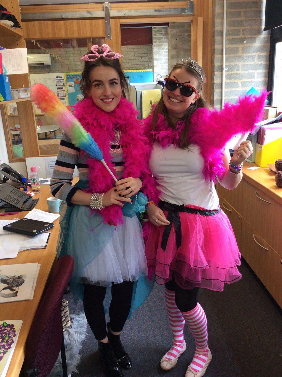 Two Women are pictured wearing a white and a pink tutu. Both have hot pink feather boas. One is holding a rainbow duster and wearing a pink crown.