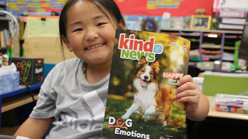 A young girl in her classroom holding up a dog magazine from kind news.