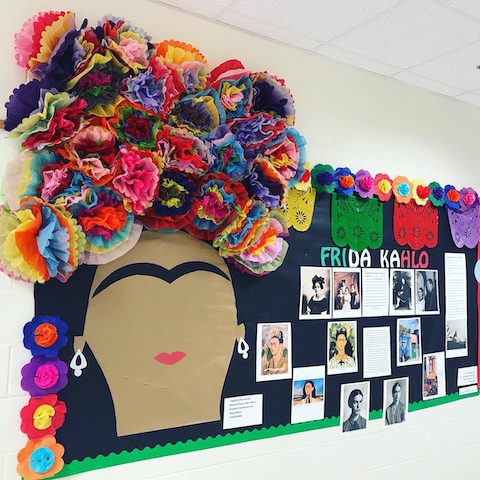 The left side of a bulletin board has a large outline of a face with a unibrow and lips as the only features. The headpiece is created from multi-color, 3-d tissue paper flowers. Various photos of Frida Kahlo are included. 