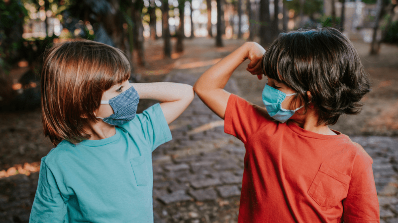 Two young boys touching elbows in the woods with their face masks on.