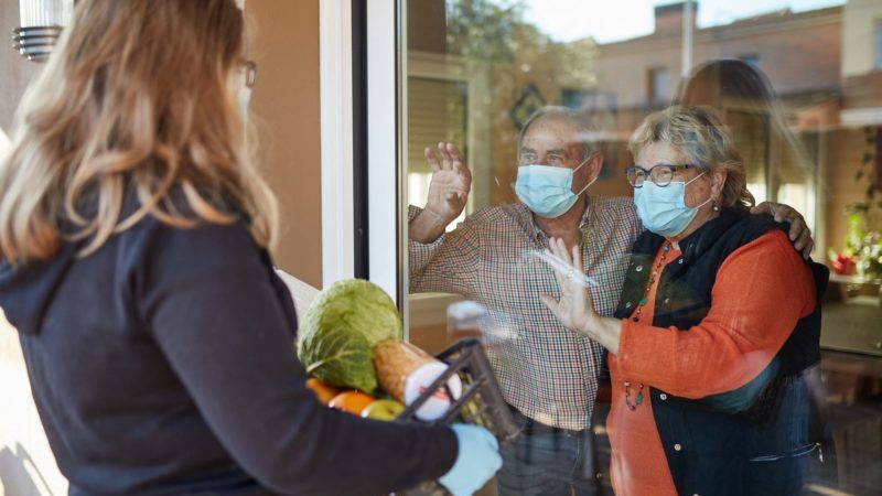 Blonde teen holds groceries in bag for older couple in mask behind glass doorVolunteer Projects Help Teens