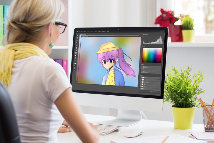 A woman sits in front of a computer that has an animated girl on the monitor with pink hair.