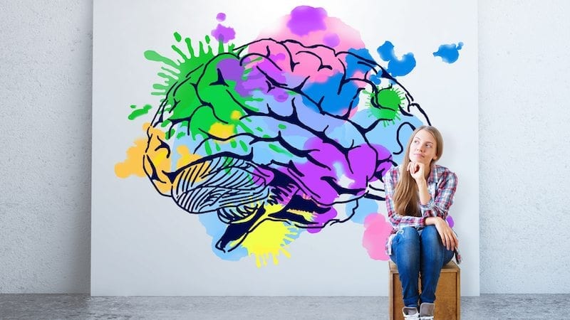 Young woman sitting in concrete room with colorful brain sketch on banner. Creative thinking concept.