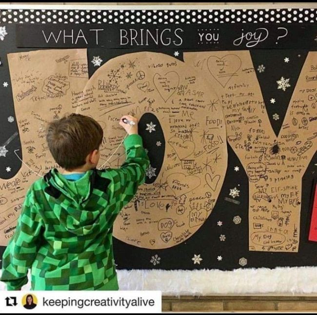 Bulletin board with large kraft paper letters spelling J-O-Y and student writing something on the letter O