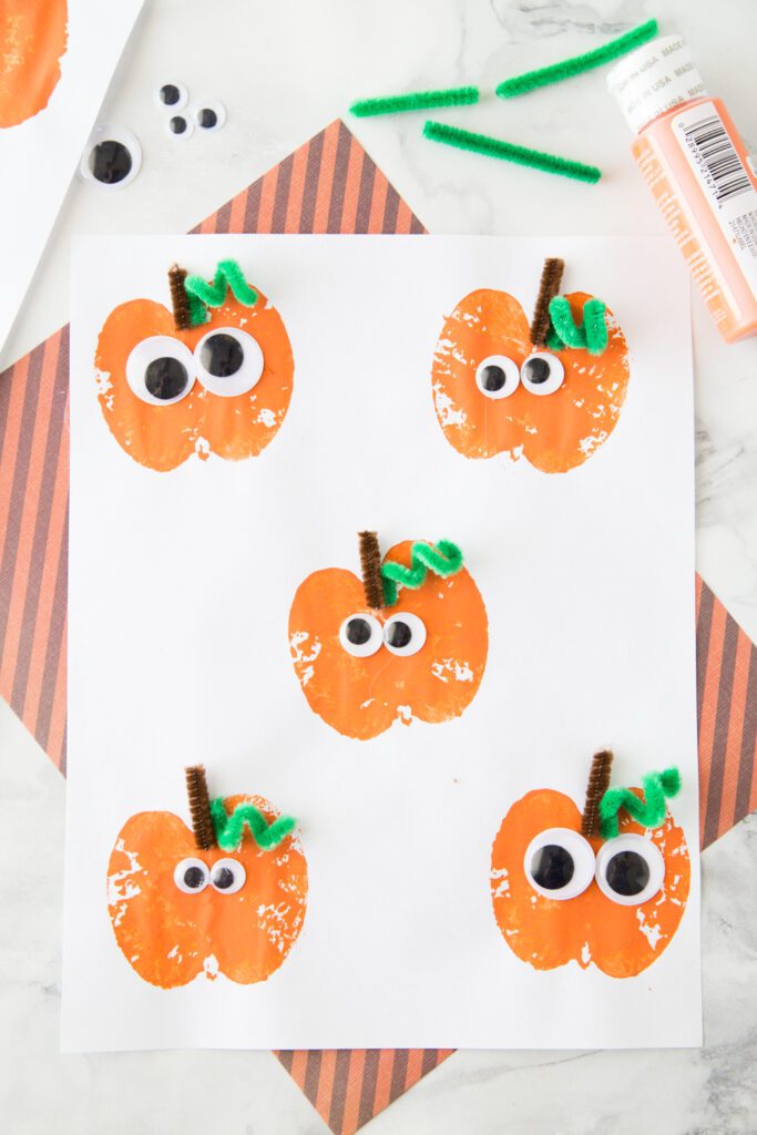 Apples sliced and dipped in orange paint are used as stamps to create pumpkin shapes on white paper. Google eyes and pipe cleaners are added.