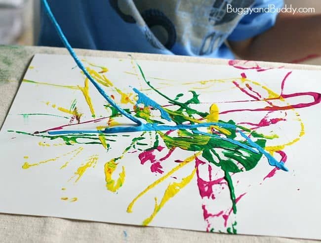 Child dragging a piece of paint-covered yarn across a piece of paper spattered with with colorful paint