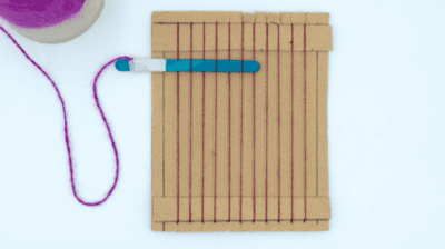 a piece of cardboard with strings strung in rows vertically and a popsicle stick with yarn tied to the end woven into the top section of the strings