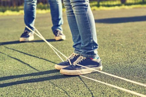 two pairs of legs, wearing jeans and tennis shoes, with a rope around one person's ankle stretching over and under the other person's feet and beyond