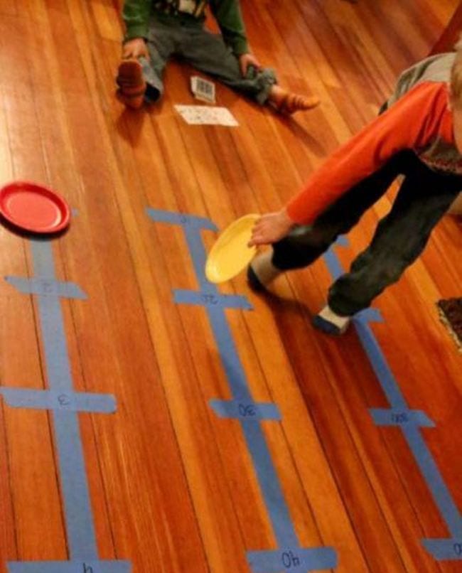  Estudiante joven moviendo platos de papel de colores a lo largo de las líneas de números hechos de cinta de pintor azul en el suelo