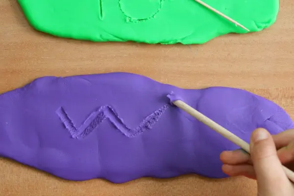 A hand is shown practicing writing with a wooden stick in play dough.
