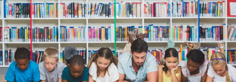 Teacher and students laying on ground in front of bookshelves reading on tablets - digital libraris