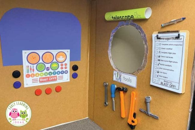 a play astronaut training center made from two sides of a cardboard box with colorful printouts attached, plus tools and a clipboard