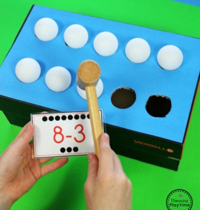 a shoe box covered on top with blue construction paper. Five holes cut into the box hold ping pong balls. A student's hands hold a wooden mallet and a card with a subtraction problem