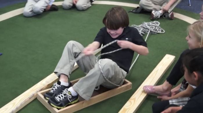 Third grade science student sitting in a wood tray, pulling himself along with a rope