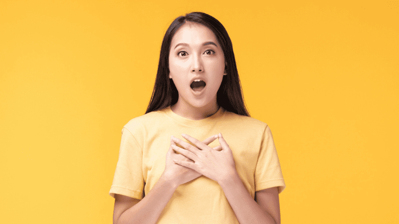 Asian woman with a shocked expression on her face standing in front of a bright yellow background