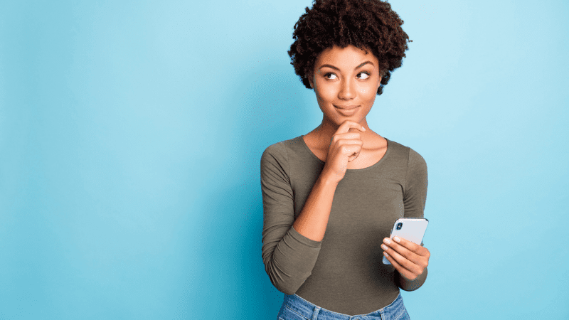 Black woman holding cell phone smirking in front of a light blue background