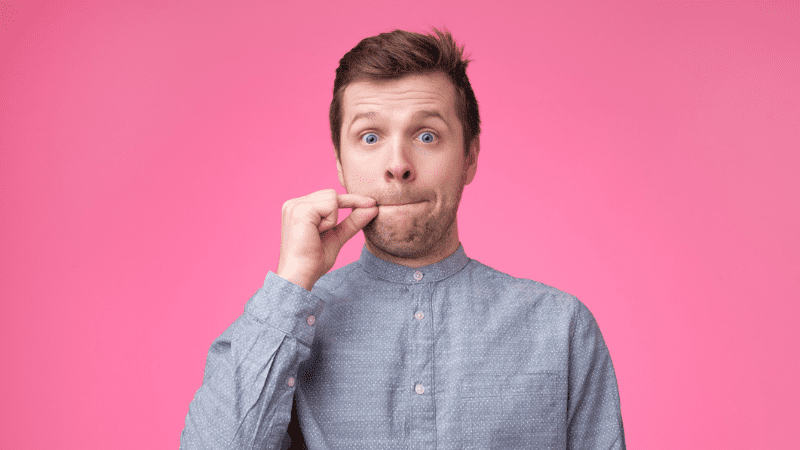 White man making the "lips sealed" sign with his mouth and fingers in front of a bright pink background