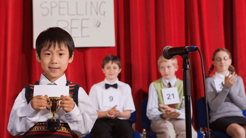 Photo of boy spelling word at a spelling be with three students sitting behind him