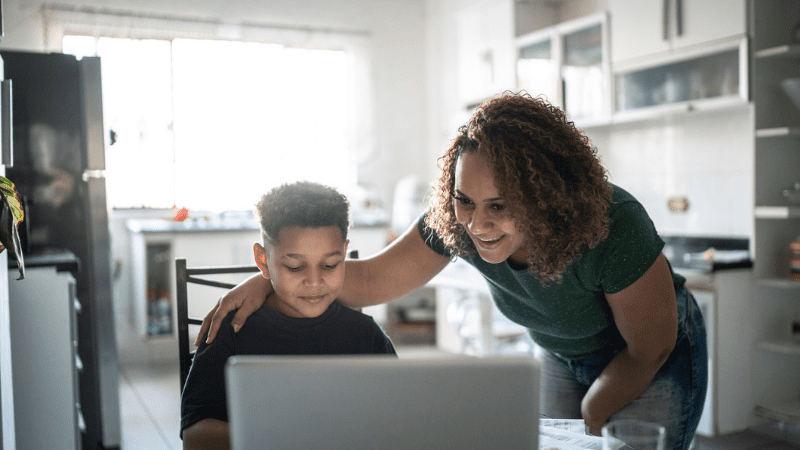 Mother helping young son as he works with virtual teachers on his laptop while sitting in his kitchen