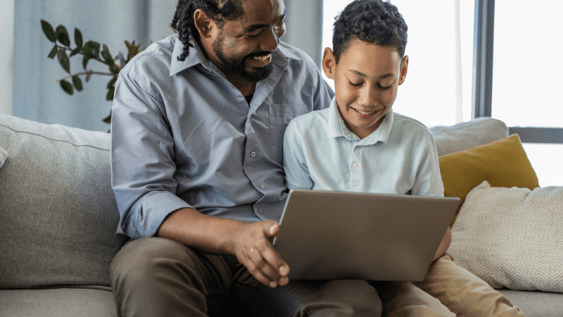 Photo of a Black father and son sitting on a couch smiling as they look at a laptop