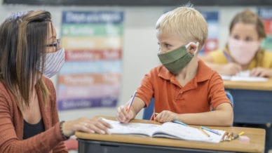 School teacher assisting student in classroom while both wearing masks to protect from the transfer of germs.