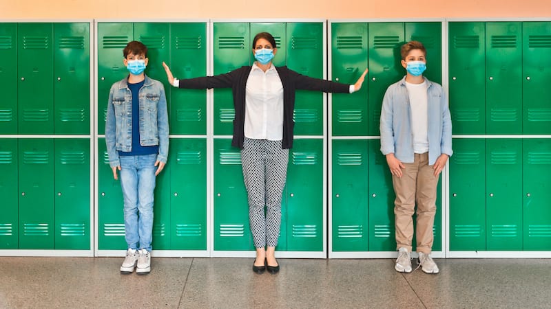 Teenage boys and female teacher wearing N95 face masks standing in front of school lockers. Woman outstretching arms in the distance.
