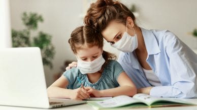 Exhausted woman in medical mask sitting at table and watching diligent girl writing in notebook while doing homework together during pandemic.