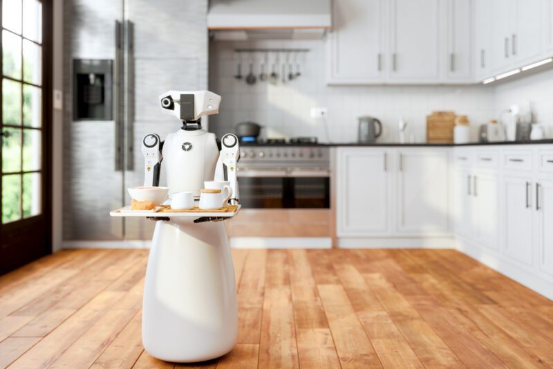 Robot Maid Holding A Tray And Serving Food And Drink In Modern Domestic Kitchen With Blurred Background. Artificial Intelligence And Smart Robotics Concept.