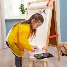 A little girl is shown mixing paints in front of an easel while wearing a yellow smock to protect her clothes.