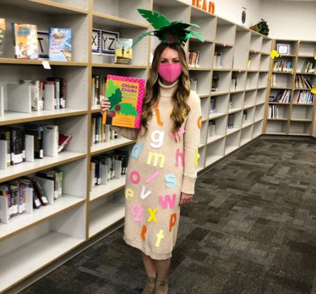 Woman wearing palm leaves on her head and covered in alphabet letters, carrying the book Chicka Chicka Boom Boom