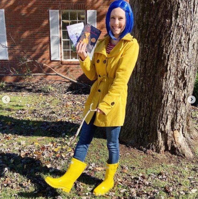 Woman wearing blue wig, yellow raincoat and yellow rainboots holding a copy of the book Coraline