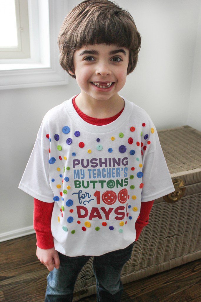A little boy is wearing a white t-shirt that says Pushing His Teacher's Buttons for 100 Days (100th day of school shirt ideas)