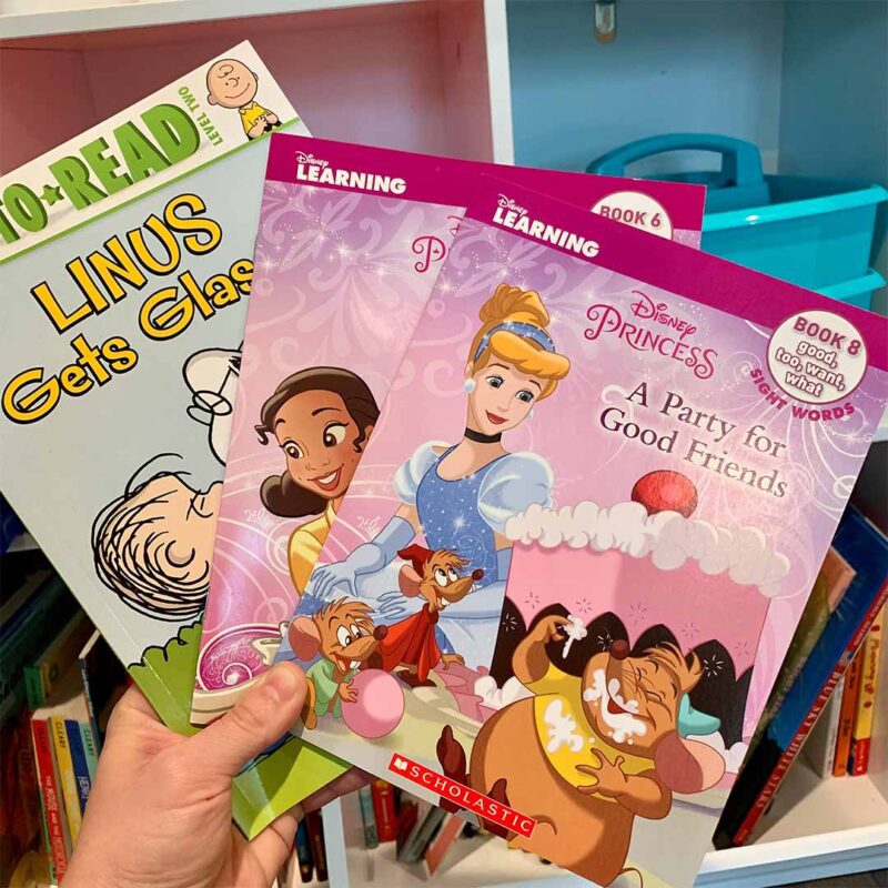 Hand holding dollar books from Books by the Bushel including Linus Gets Glasses and two Disney princess easy reader books.