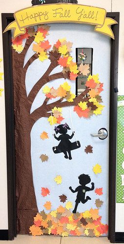 A classroom door is decorated with the side of a tree with fall leaves. A swing has a silhouette of a little girl on it swinging from a branch. A silhouette of a little boy is shown playing in the leaves beneath.