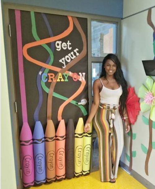 Teacher standing by a classroom door decorated with 3D paper crayons. Text reads 