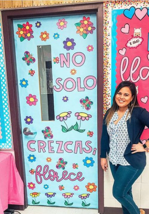 Teacher standing next to a classroom door decorated with paper flowers. Text reads No solo crezcas, florece.