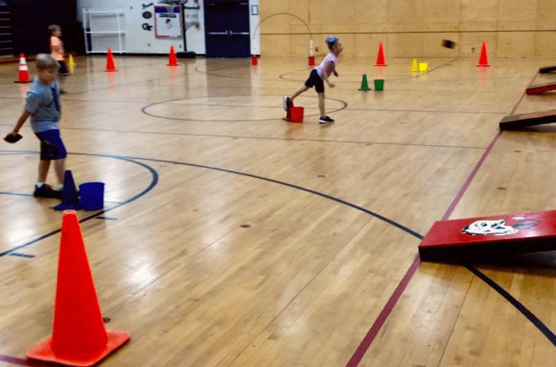 Students stand about 10 yards back from cornhold boards. There are cones scattered throughout the gymnasium.