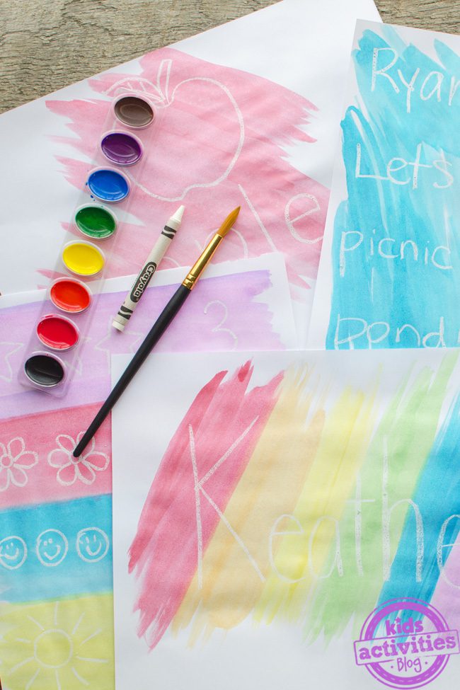 This easy art project for kids shows multiple pieces of paper with different doodles and words in white crayon with paint surrounding it. An actual white crayon and watercolor paints are on top of the paintings.